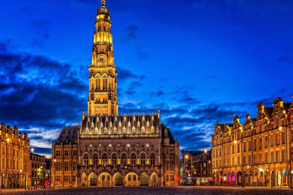 Le beffroi d'Arras illuminé à la nuit tombée, entouré par les bâtiments historiques de la Grand-Place.